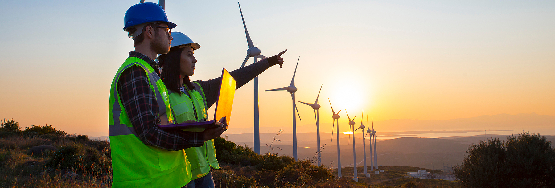 Section Image: Two people standing on a ridge holding a laptop with a row of windmills 