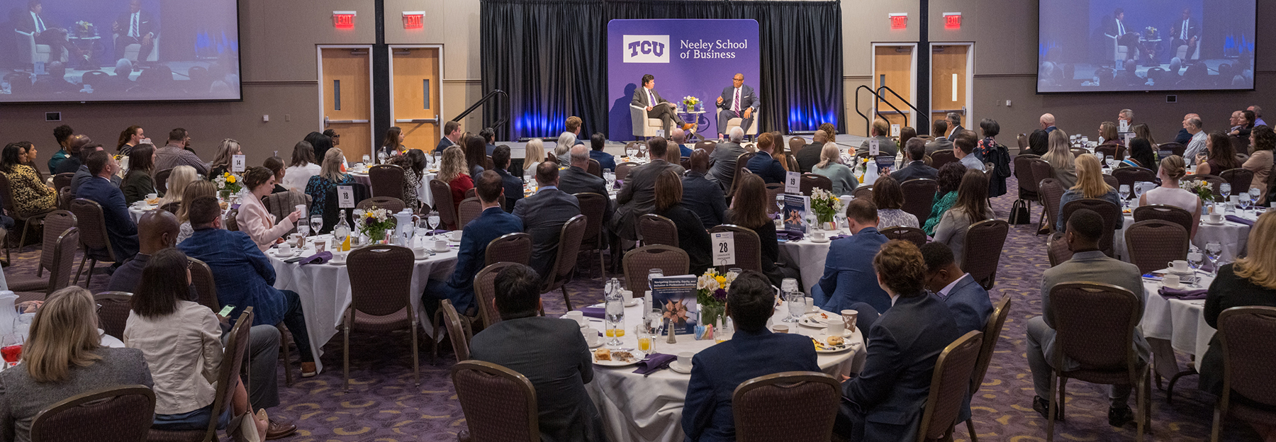 Section Image: Dean Pullin and Key Bouyer on the stage in front of a room full of people at round tables eating breakfast. 