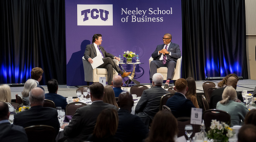 Section Image: Dean Pullin and Key Bouyer on the stage in front of a room full of people at round tables eating breakfast. 