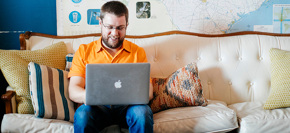 Section Image: Man sitting on a couch with a laptop and a map on the wall behind him. 