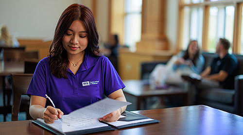 A girl in a TCU polo sitting at a desk with pad and pen.